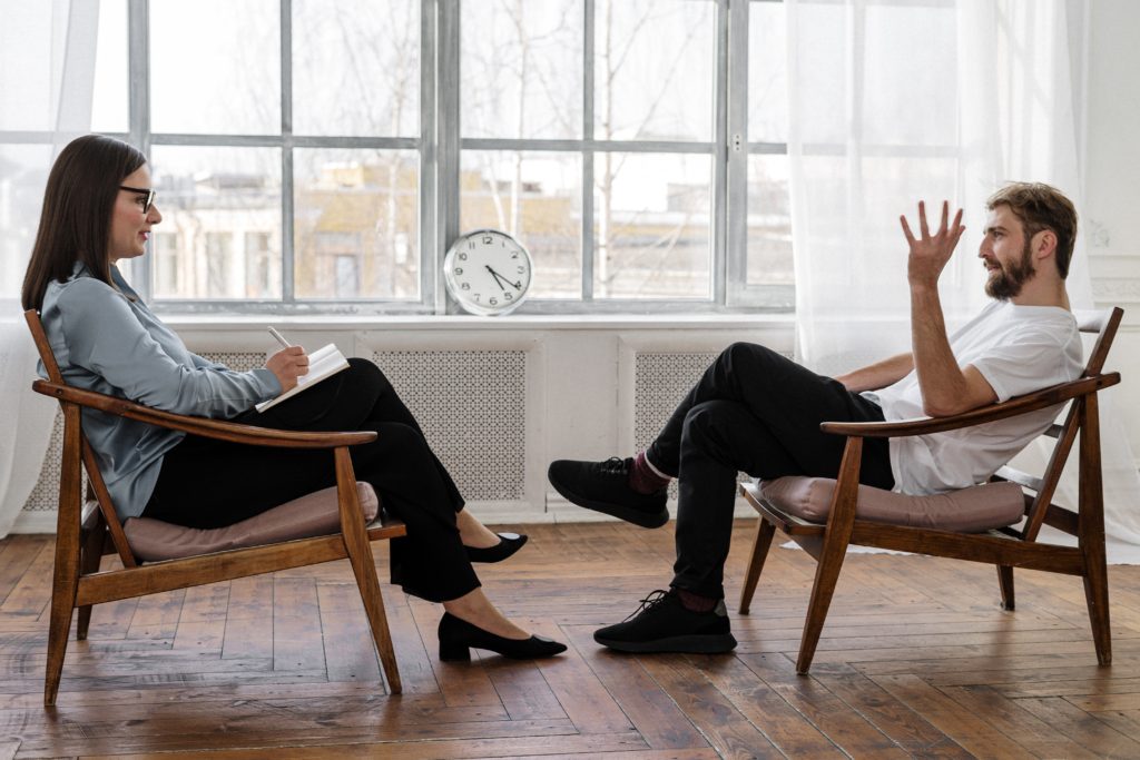 A woman and a man have a discussion while sitting in wooden chairs.